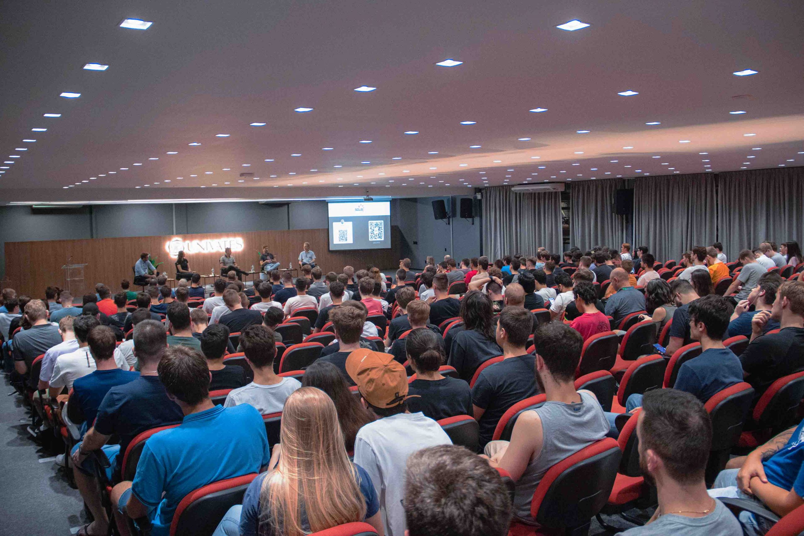 Imagem mostra diretor e painelistas em auditório da Univates, com estudantes da graduação em tecnologia na plateia